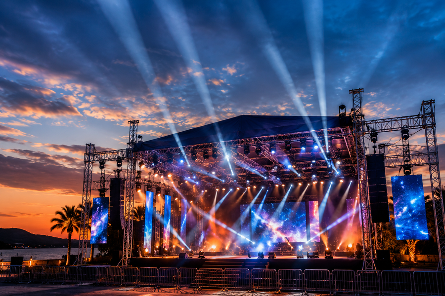 Large outdoor concert stage at sunset with powerful beam moving head lights shooting into the twilight sky, LED screens flanking the stage, and professional truss rigging silhouetted against orange and blue clouds